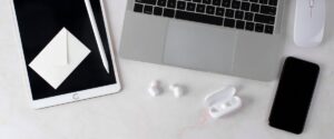 Ambiente clean e minimalista com itens de home office. Sobre uma superfície clara, estão dispostos um iPad branco com uma caneta ao lado, um envelope pequeno branco e mouse branco